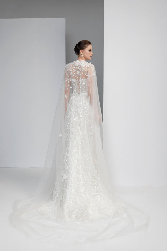 Woman wearing a white wedding dress with a long train and sheer sleeves against a plain background