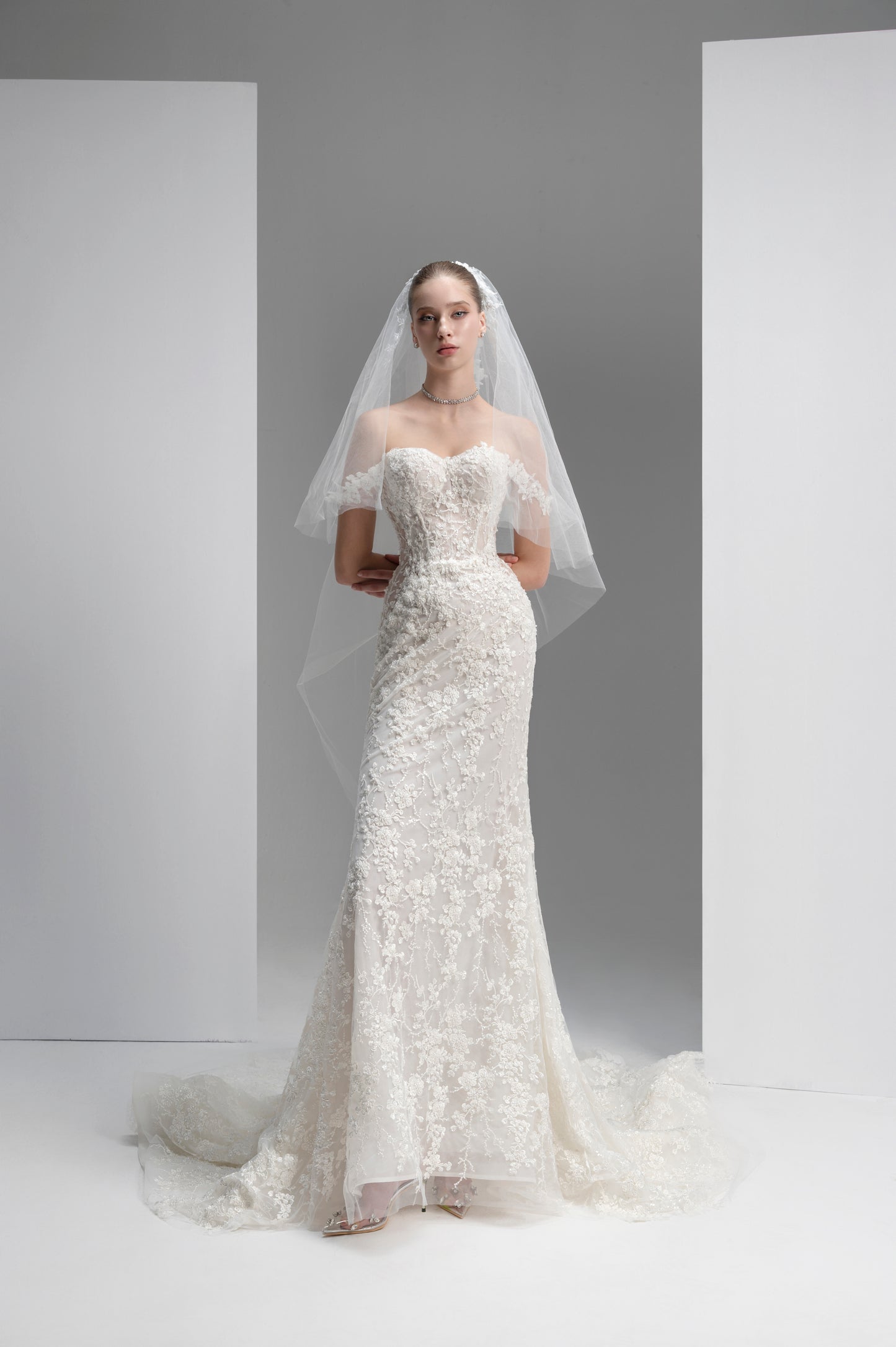Model wearing a white lace wedding dress with a veil against a plain background