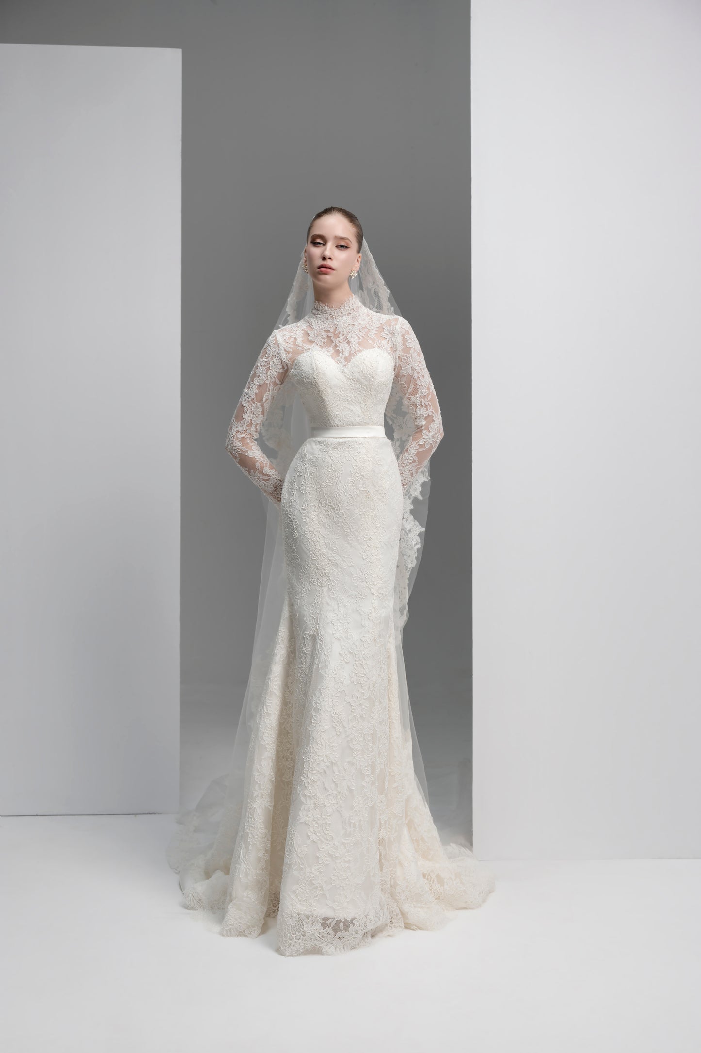 Woman wearing a white lace wedding dress with a long veil against a plain background
