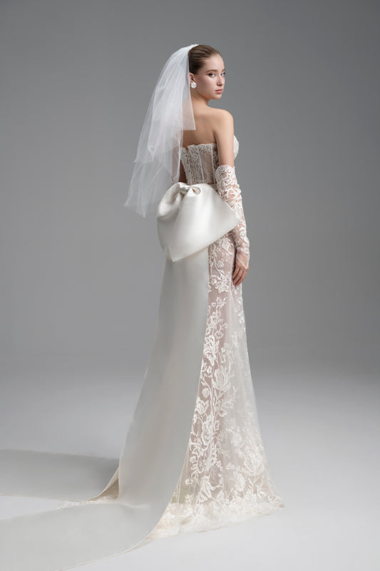 Woman in a white wedding dress with lace detailing and a veil, holding a white clutch against a gray background.