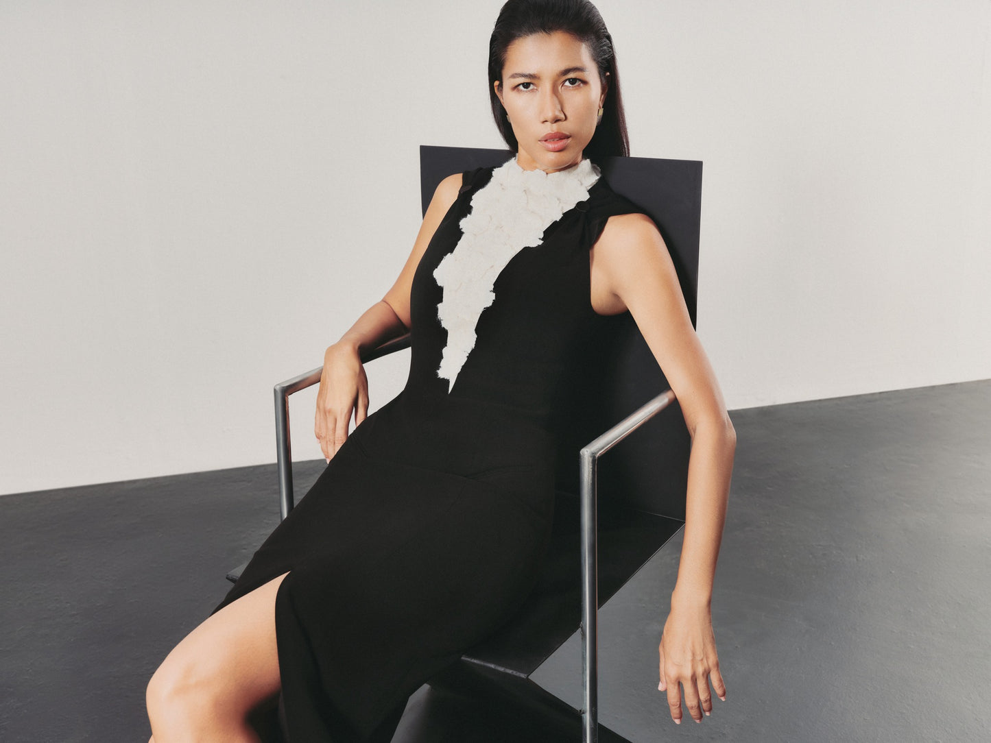 Woman in a black dress with white details sitting on a chair against a plain background