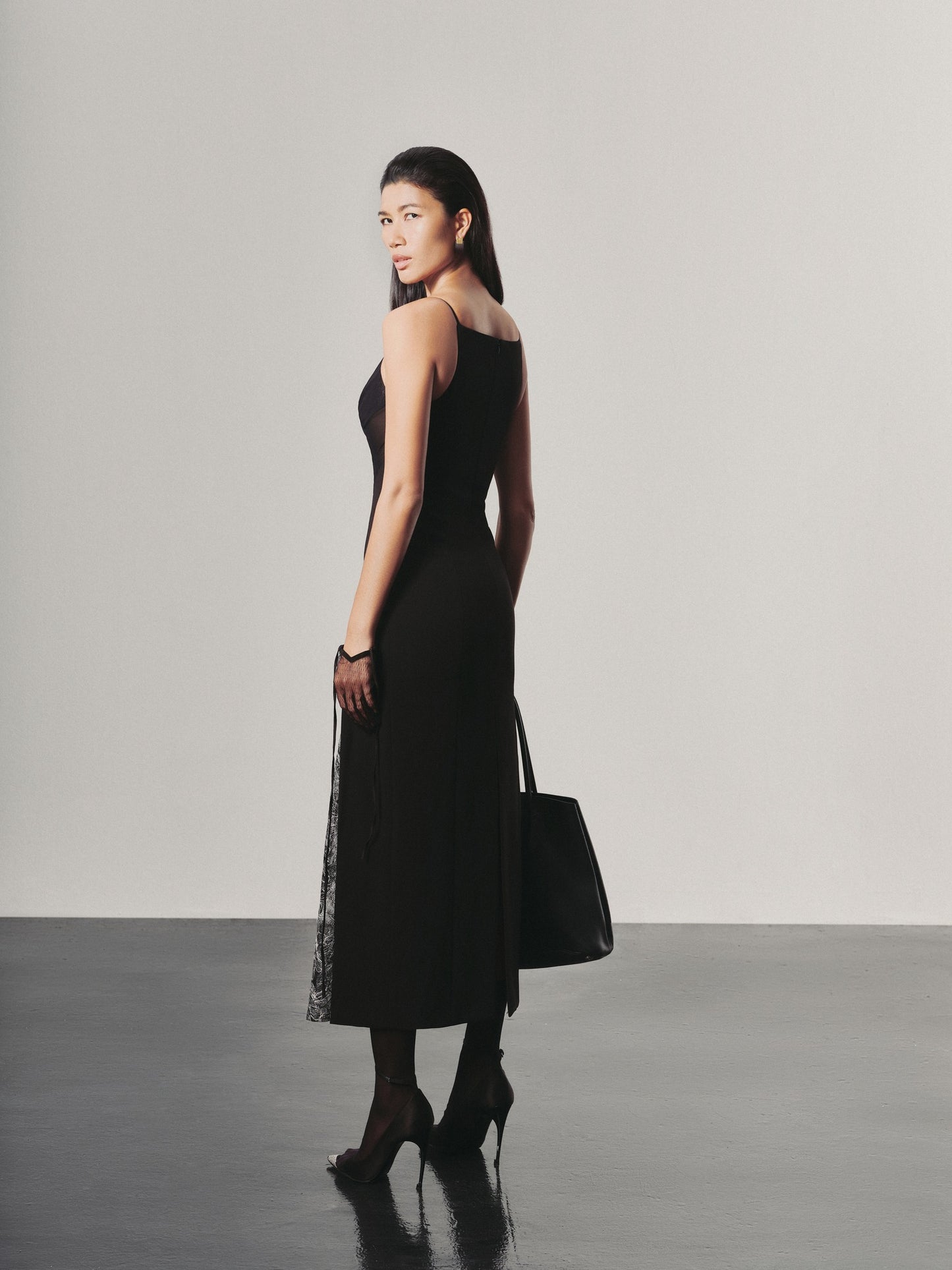 Woman in a black dress holding a black handbag against a plain background