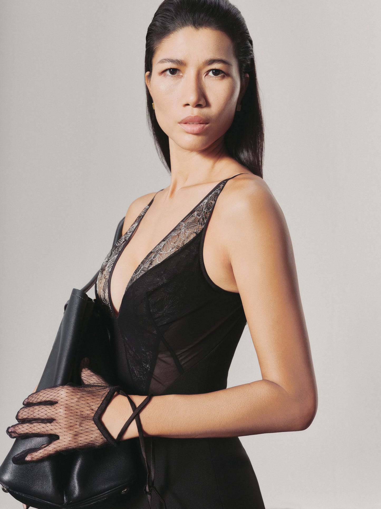 Woman wearing a black lace dress and holding a black handbag against a plain background