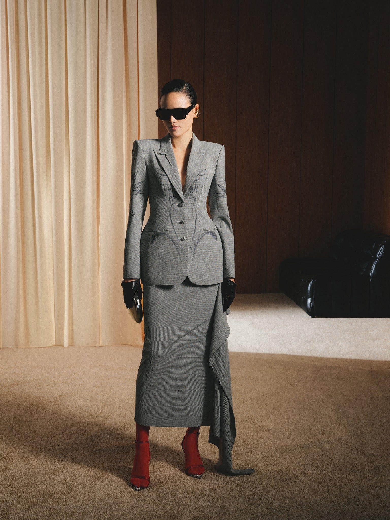Woman in a gray suit with red shoes standing in a room with beige curtains and a dark floor.