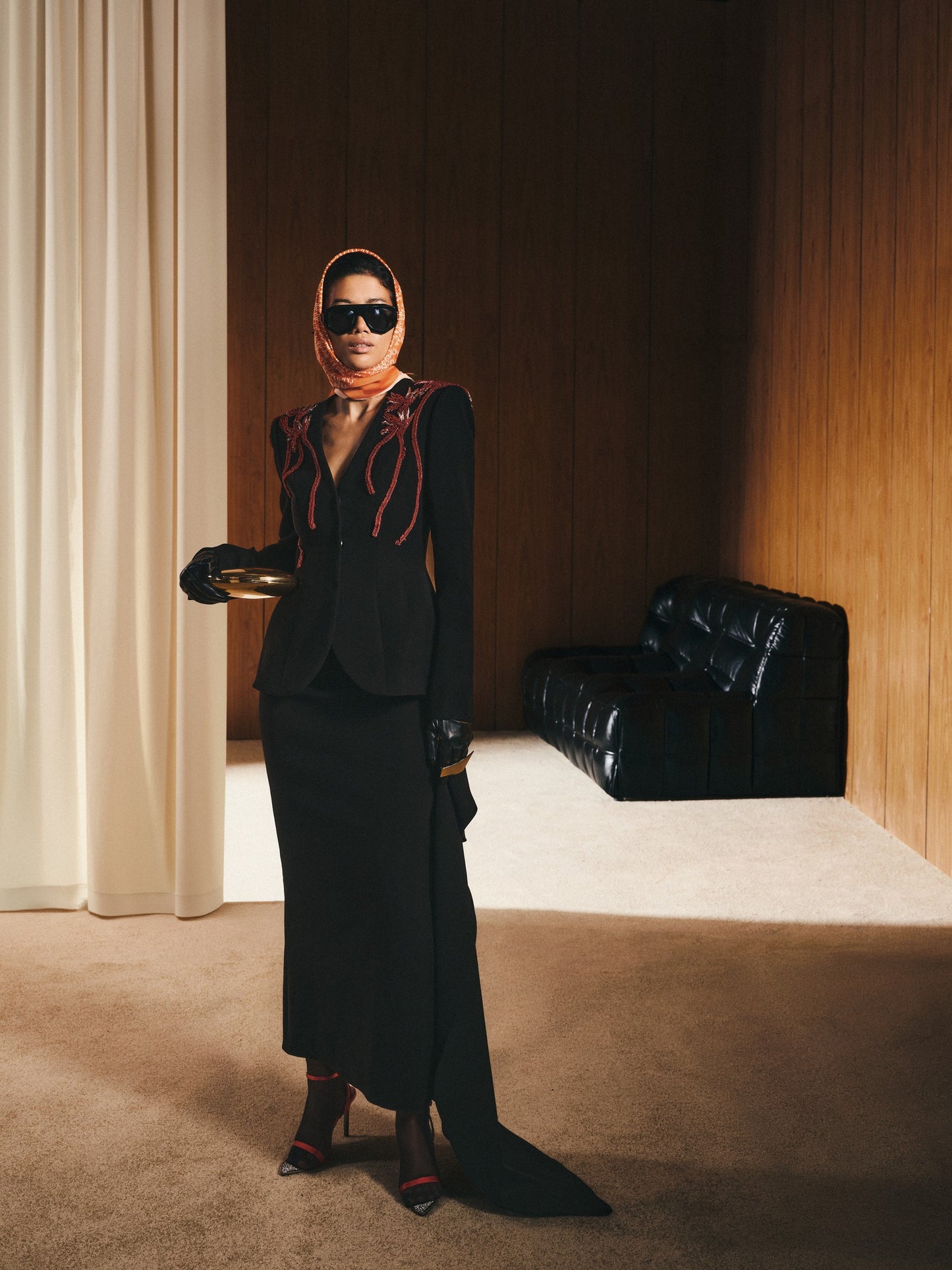 Person wearing a black outfit with a red scarf in a room with a black leather chair and white rug.