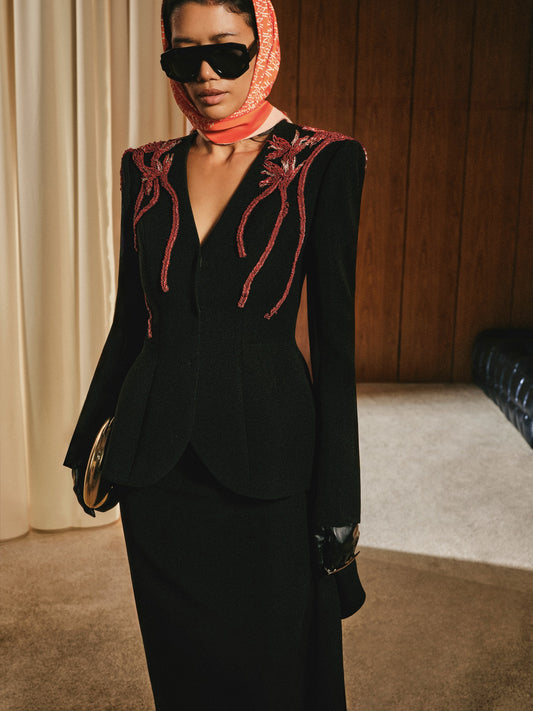 Person wearing a black outfit with red embroidery, sunglasses, and a headscarf in an indoor setting.