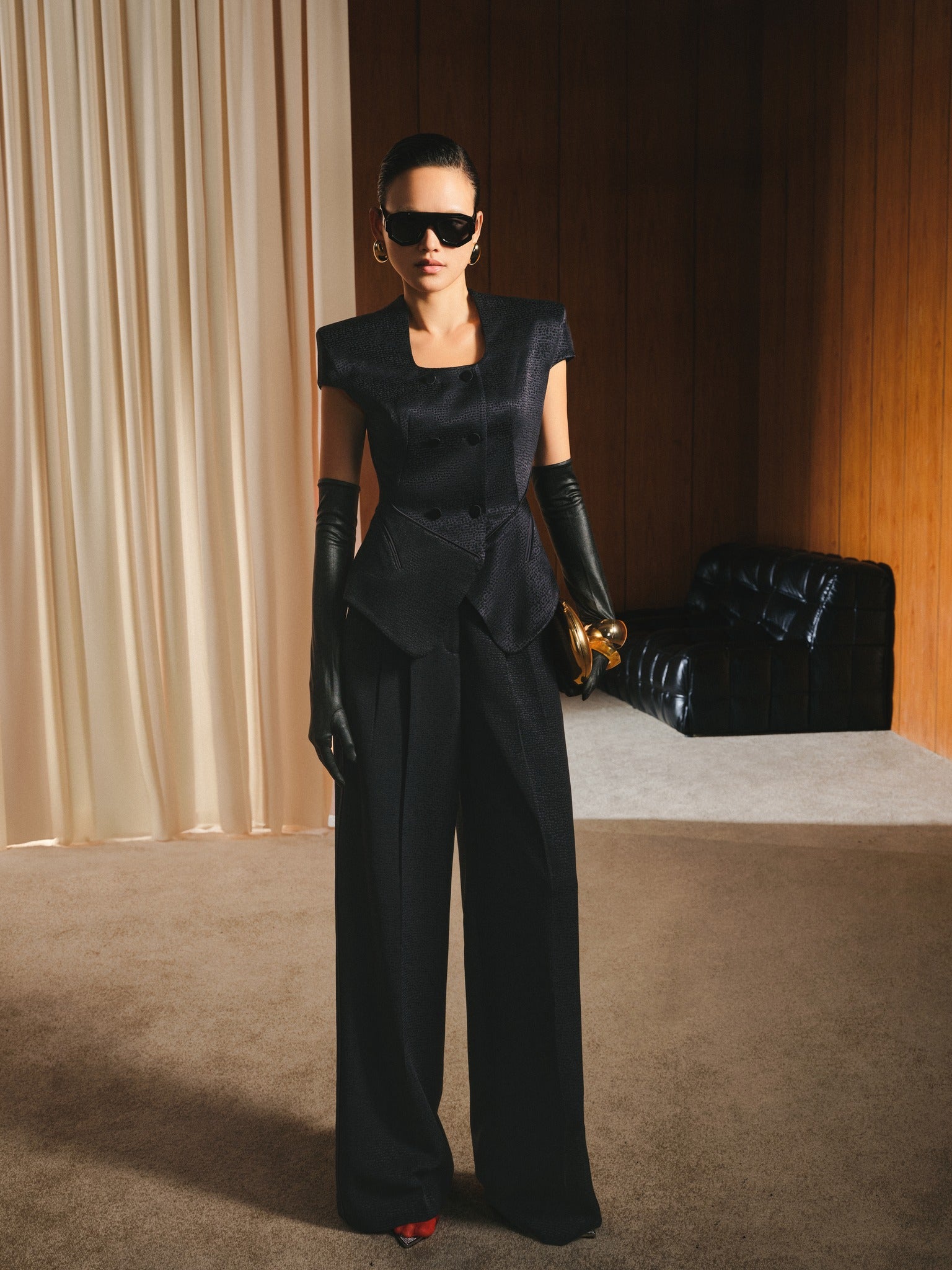 Woman in a black outfit with gloves and sunglasses standing in a room with beige curtains and a black chair.