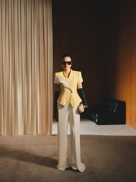 Person wearing a yellow blazer and white pants standing in a room with curtains and a black chair.