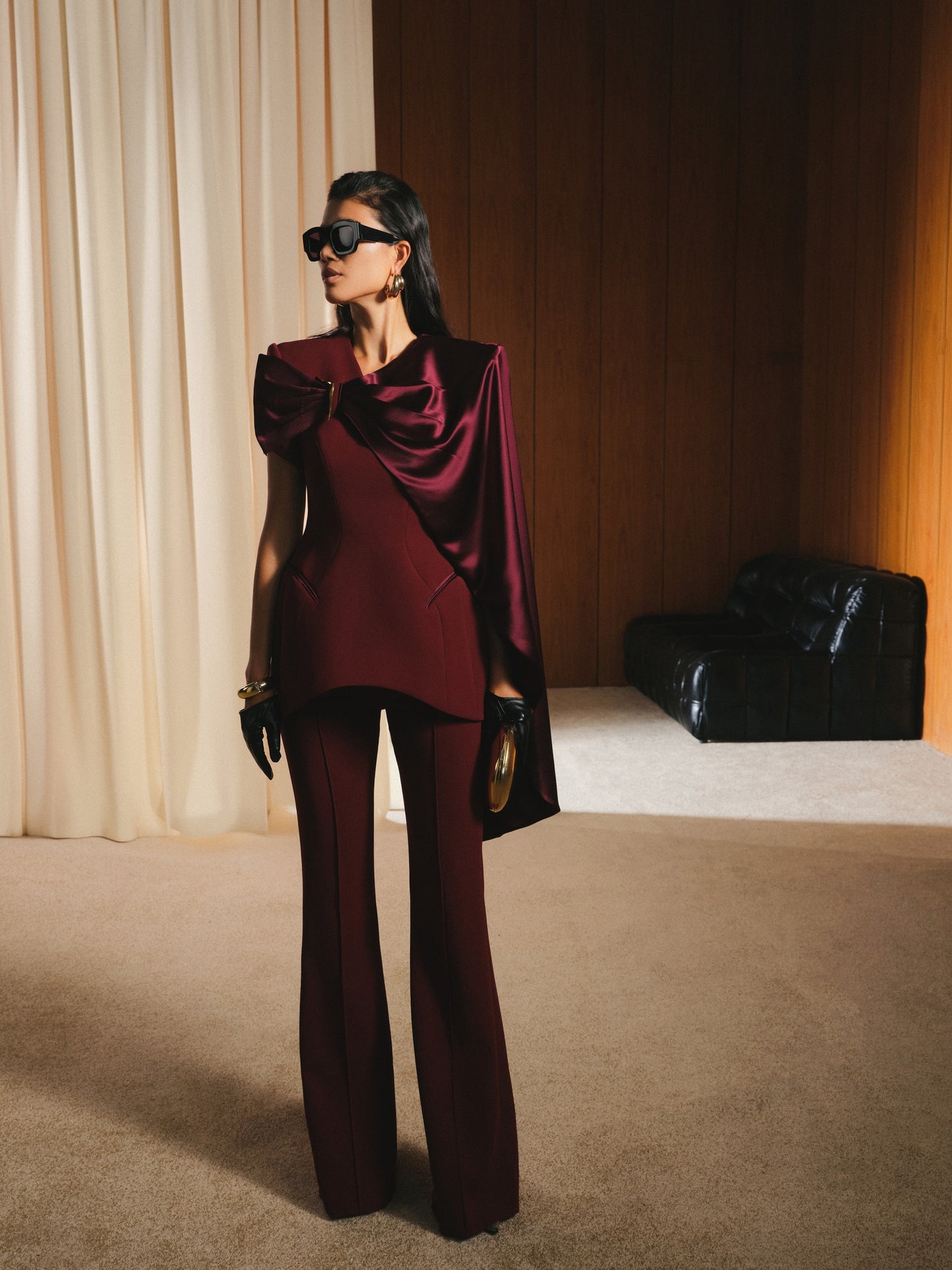 Woman in a burgundy outfit with a cape in a room with beige curtains and a black chair.