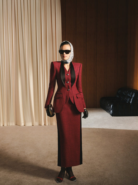 Woman in a red suit standing in a room with beige curtains and a black leather couch.