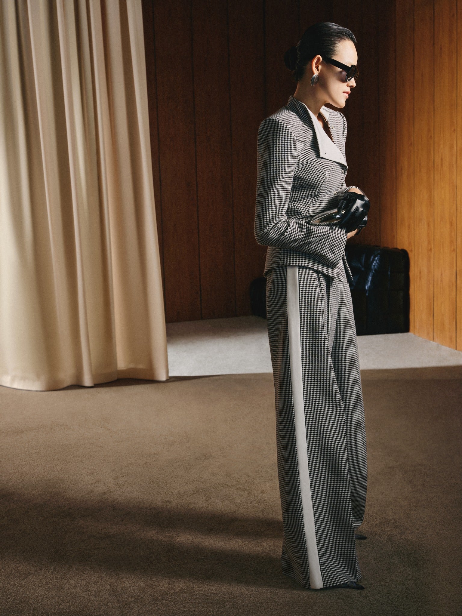 Person wearing a gray outfit with a white collar, standing in a room with beige curtains and wooden walls.