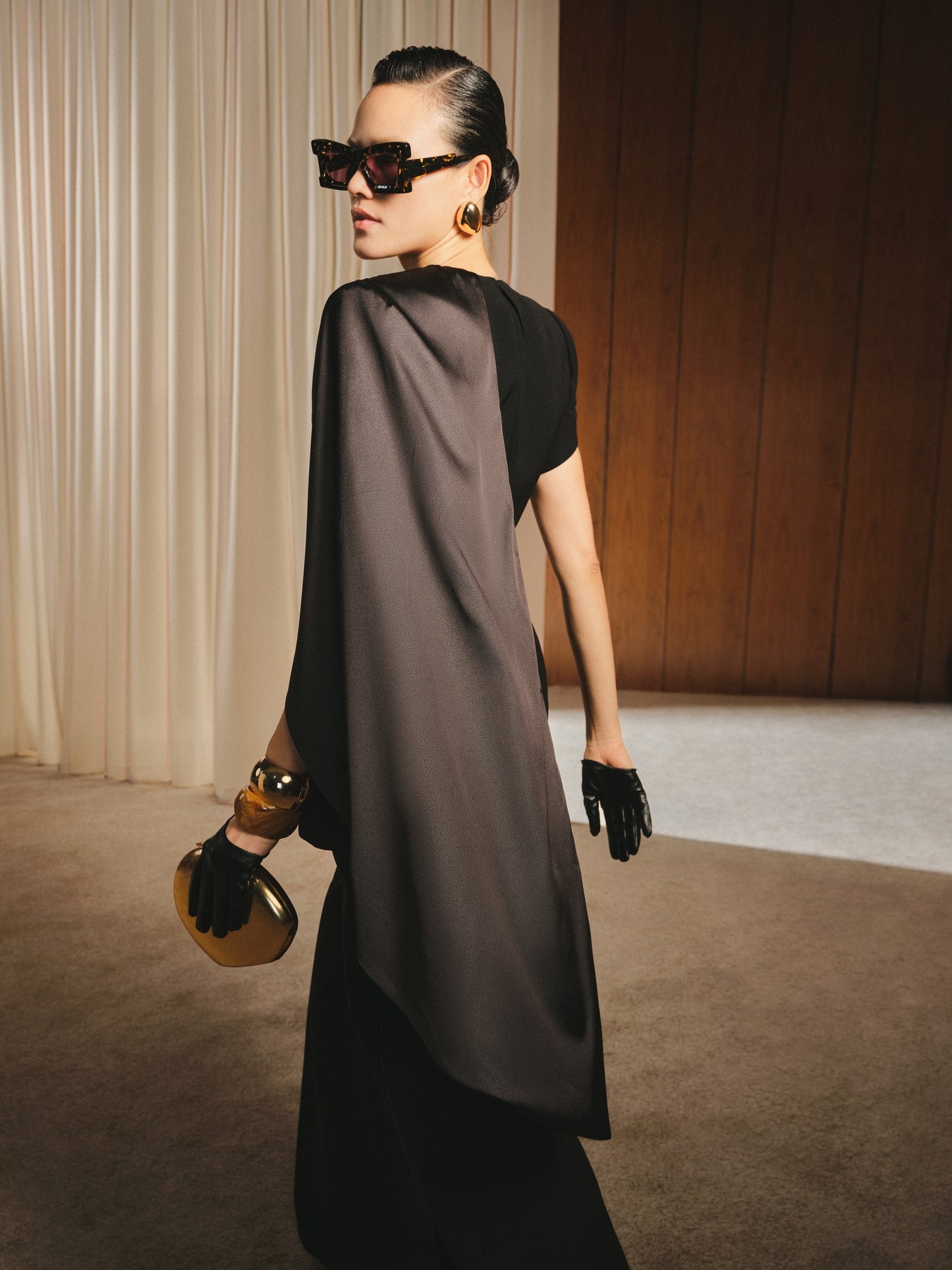 Woman in a stylish outfit with a black top and gray cape, holding a gold clutch, against a neutral background.