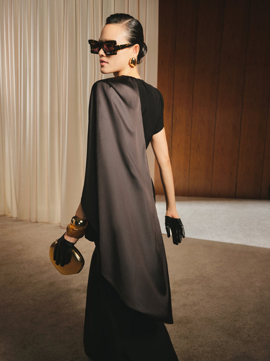 Woman in a stylish outfit with a black top and gray cape, holding a gold clutch, against a neutral background.