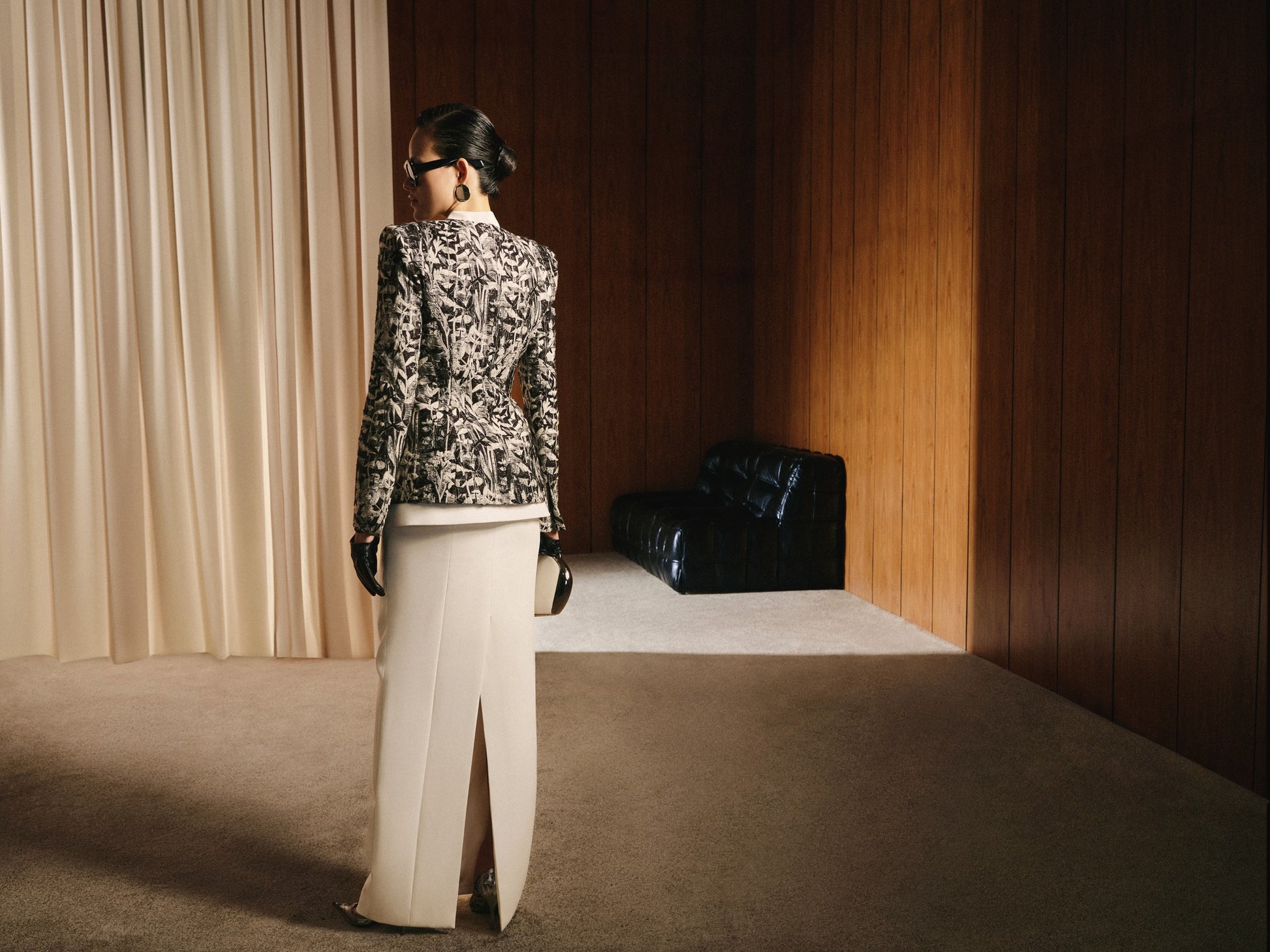 Person wearing a patterned jacket and white pants standing in a room with wooden walls and a black chair.