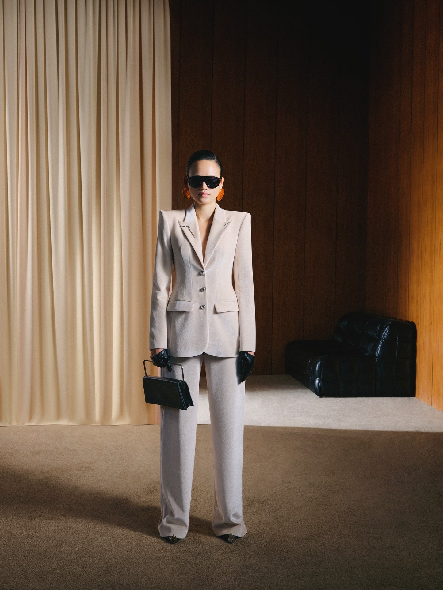 Person wearing a light gray suit standing in a room with beige curtains and a black chair.