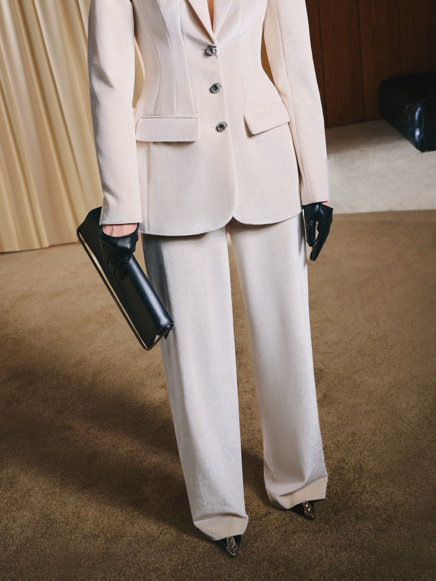 Person wearing a beige suit with black gloves and clutch in an indoor setting