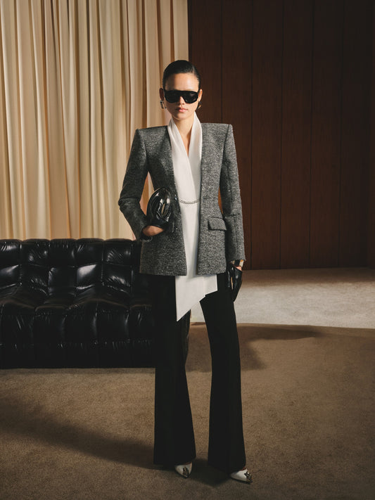 Woman in a gray blazer and black pants standing in a room with a black leather couch and beige curtains.