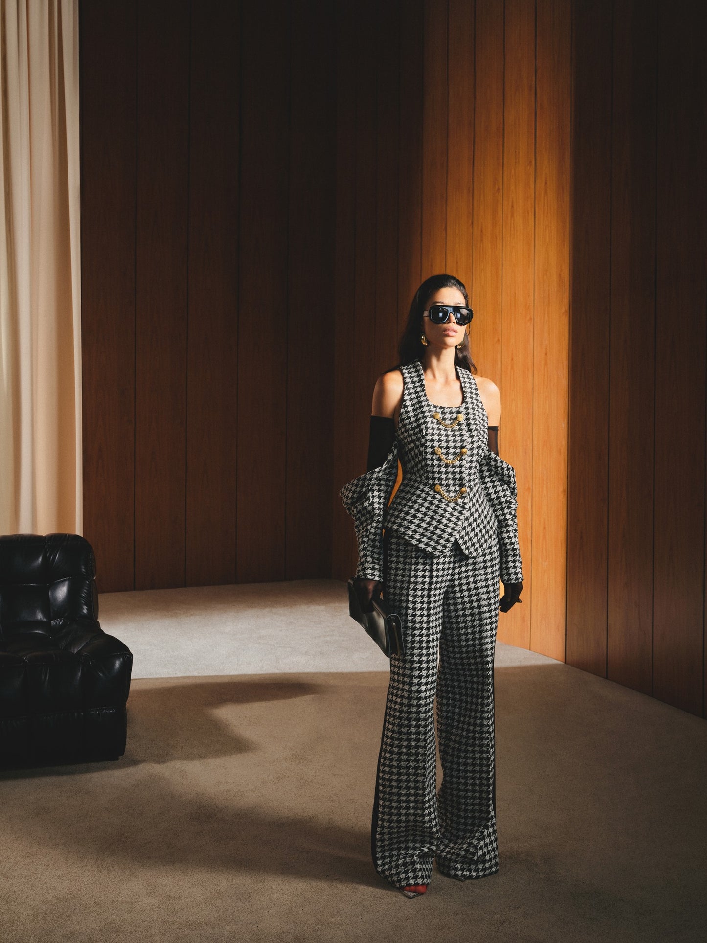 Woman in a checkered suit standing in a room with wooden walls and a black chair.