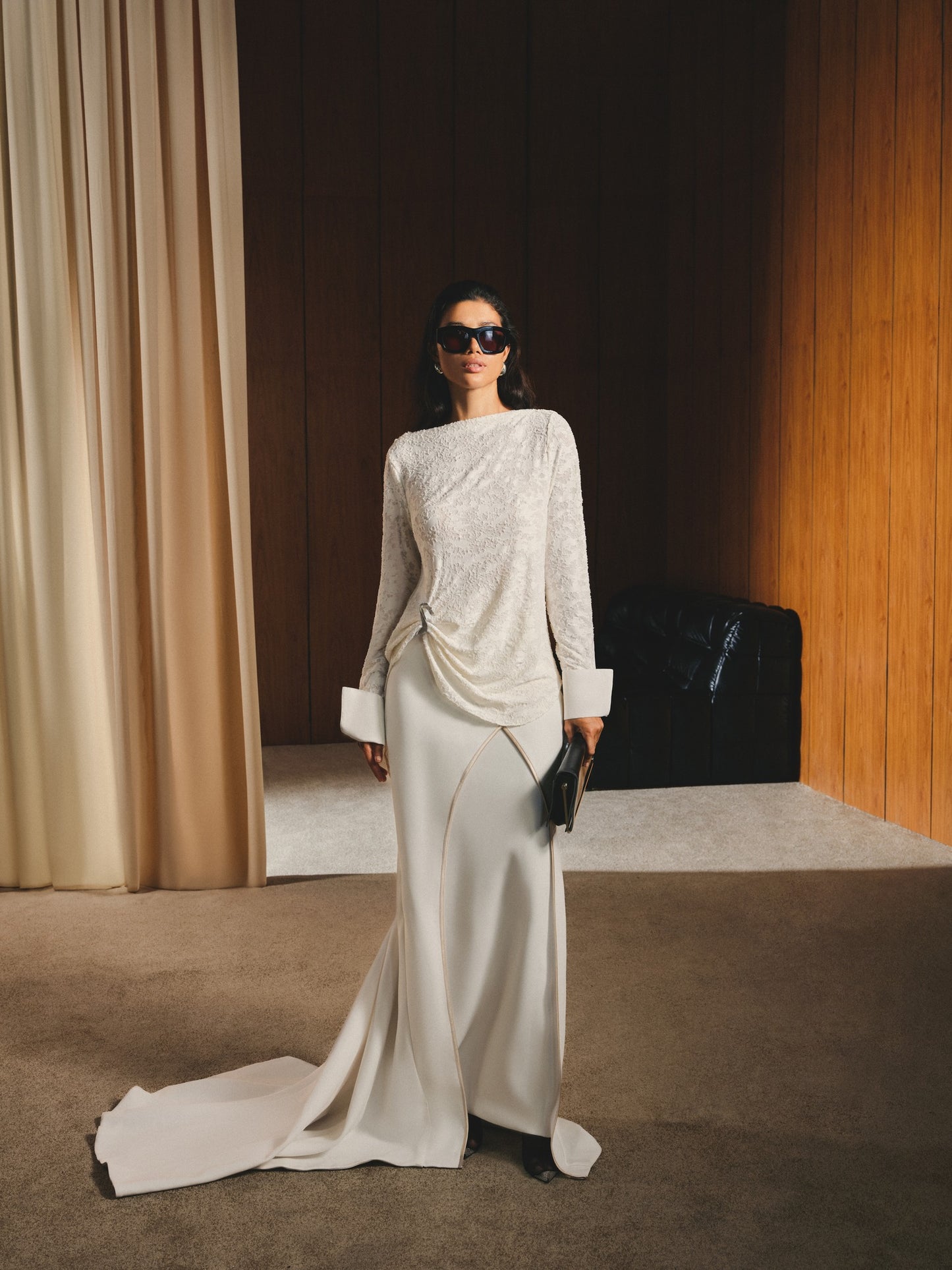 Woman in a stylish outfit standing in an elegant room with curtains and a sofa.