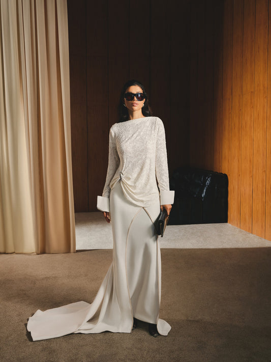 Woman in a stylish outfit standing in an elegant room with curtains and a sofa.