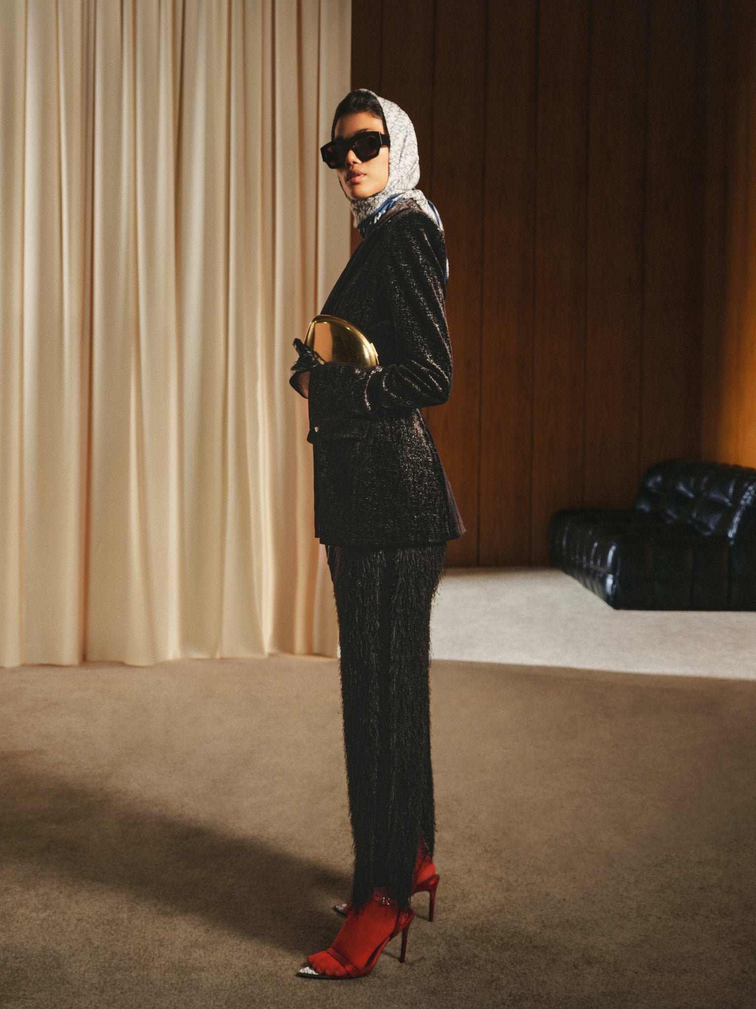 Woman in a black textured suit with red shoes standing in a room with beige curtains and a black chair.