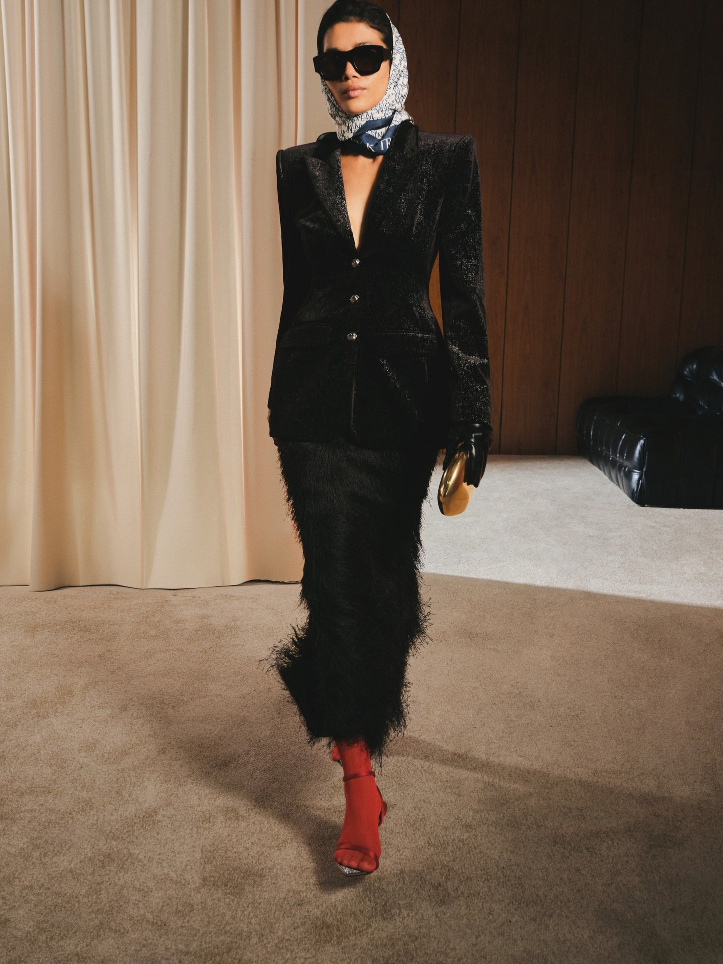 Person wearing a black suit with red shoes in a room with beige curtains and a wooden wall.