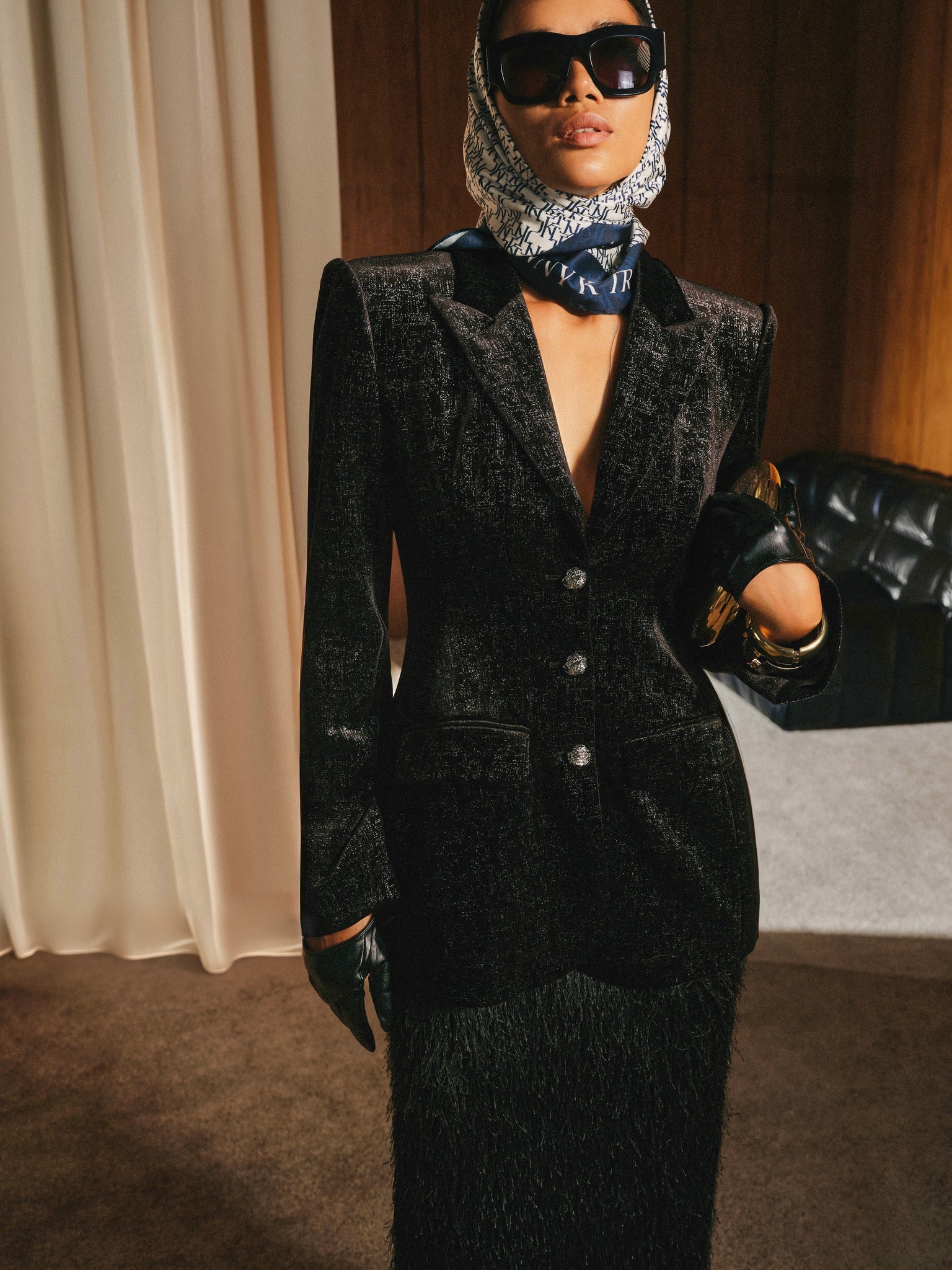 Person wearing a black textured suit with a patterned headscarf and sunglasses indoors.