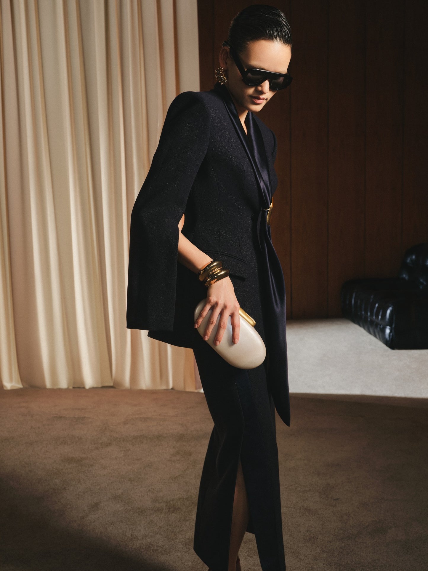Woman in a black suit holding a white clutch in a room with beige curtains.