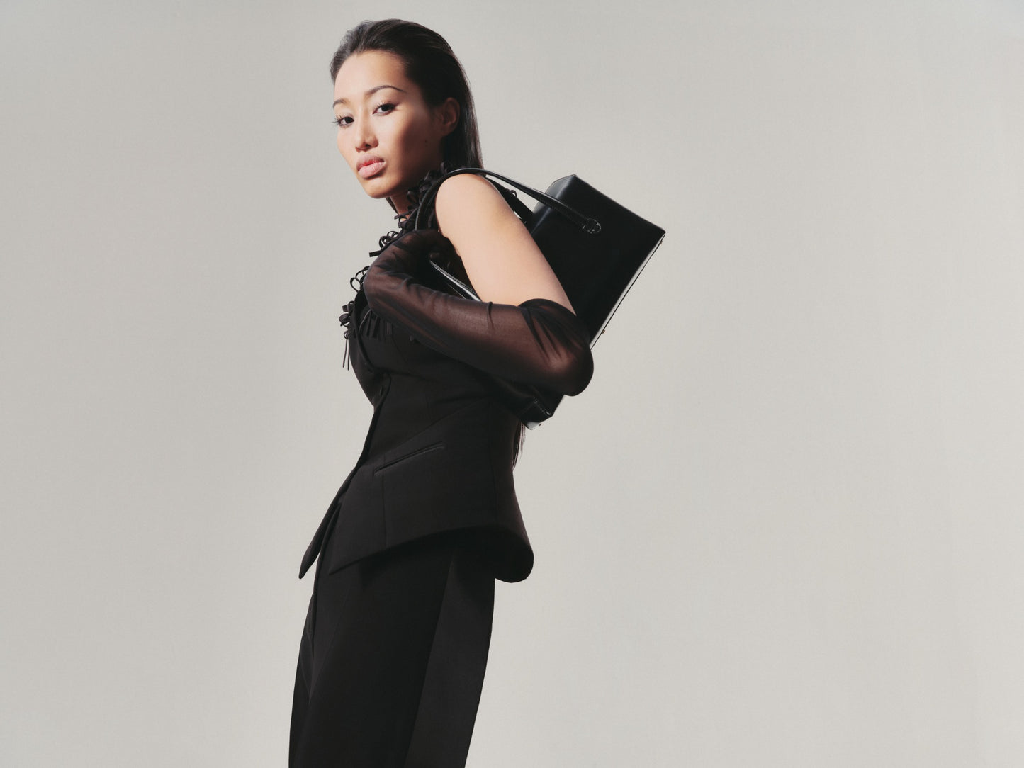 Woman in a black outfit holding a black handbag against a plain background