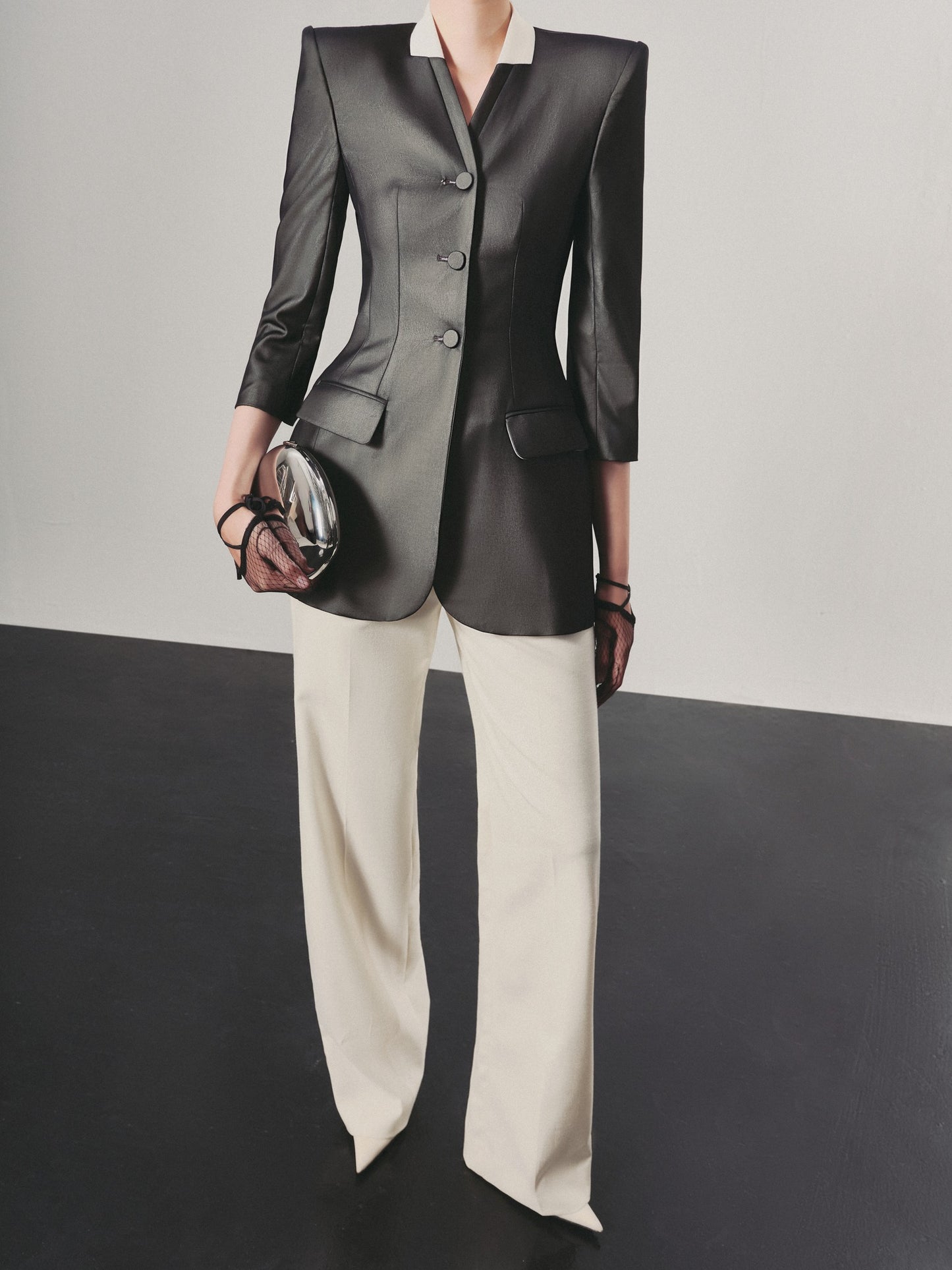 Person wearing a black blazer and white pants holding a clutch against a gray background
