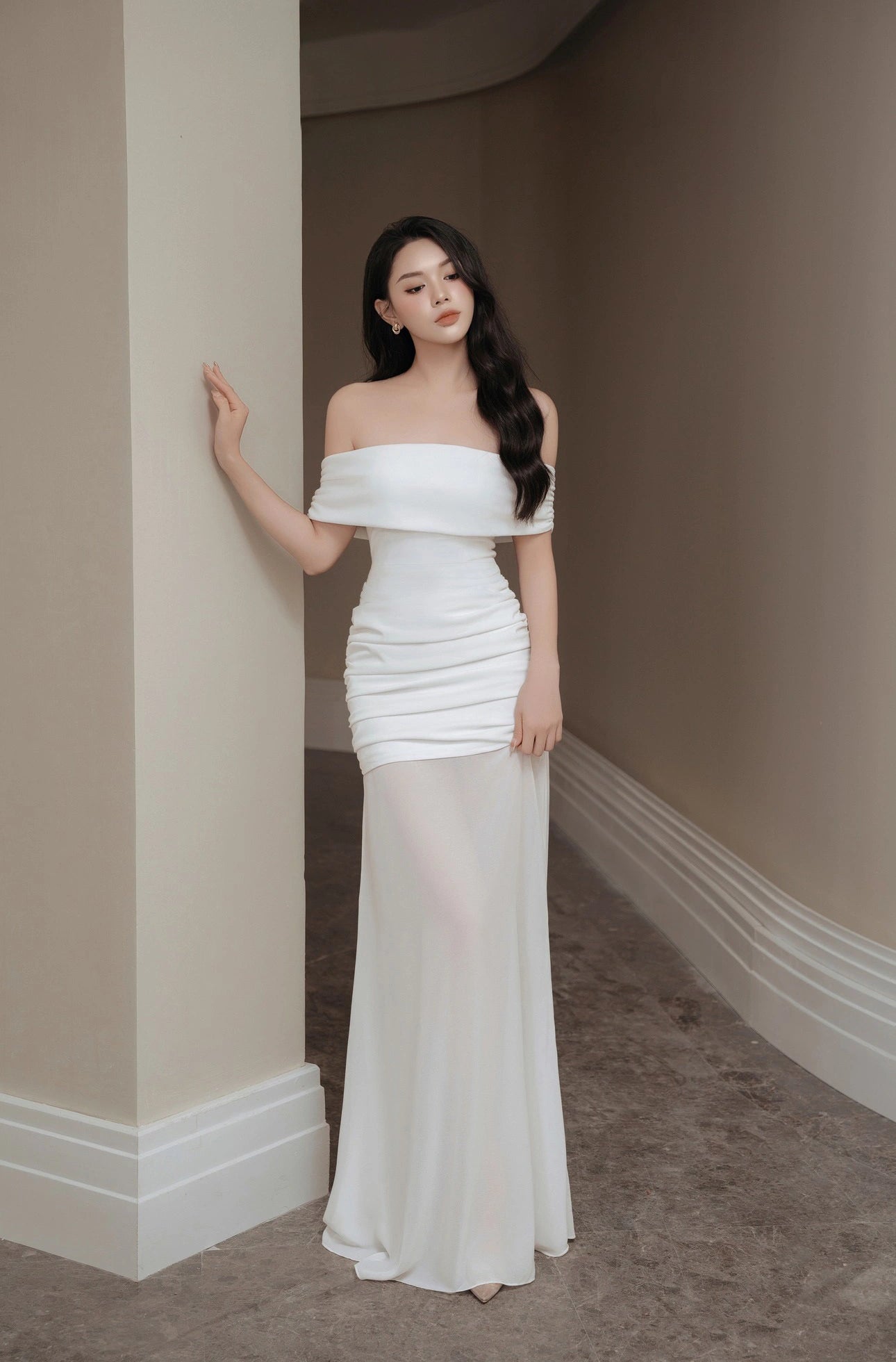 Woman in a white off-shoulder gown standing against a beige wall.