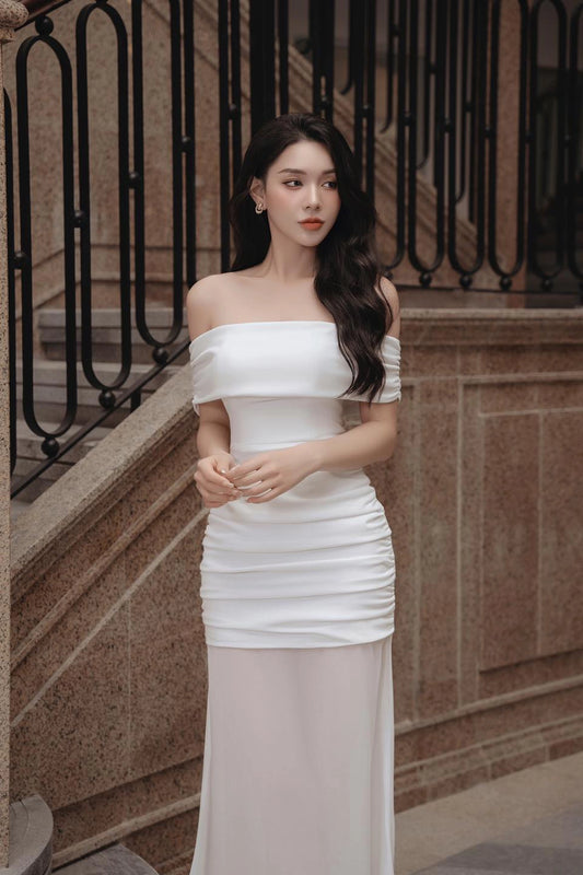 Woman in a white off-shoulder dress standing outdoors with decorative railing and wall in the background