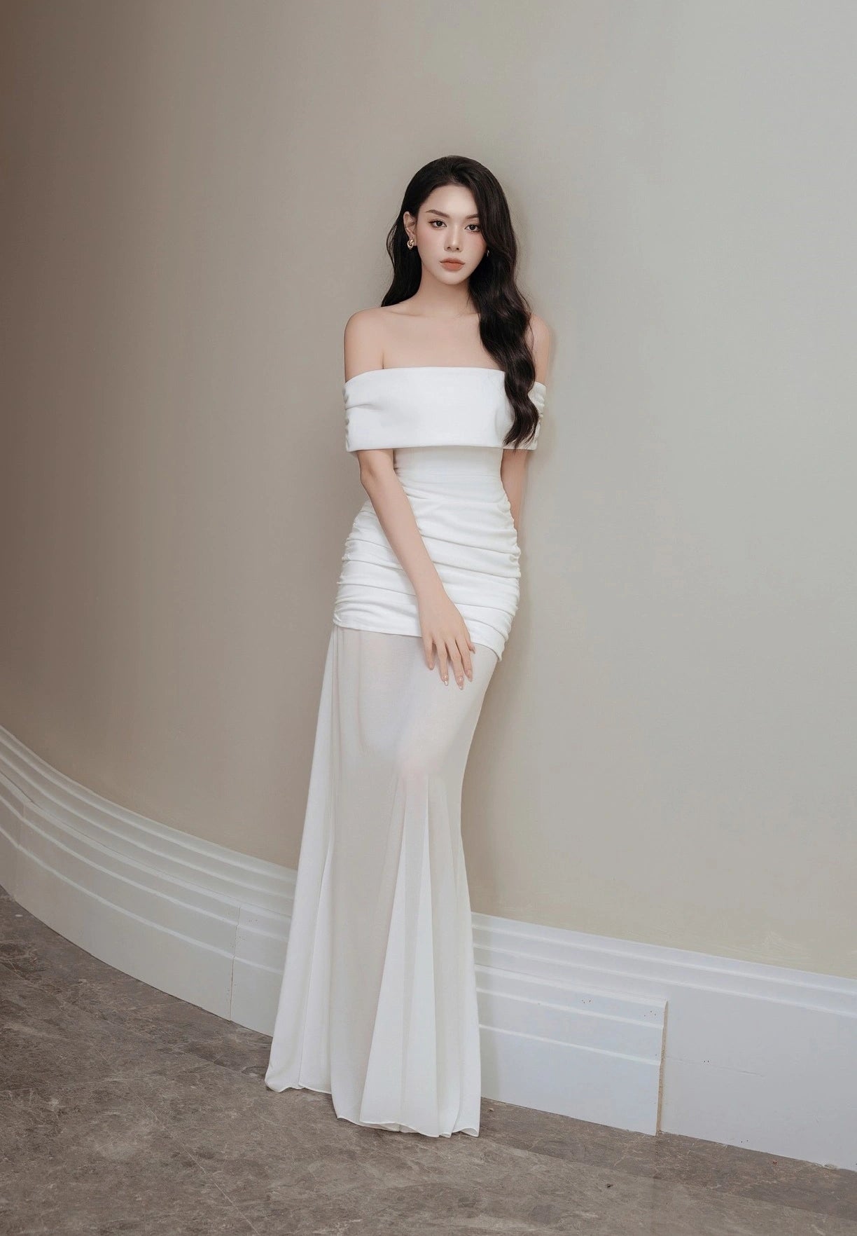 Woman wearing a white off-shoulder gown against a beige wall.
