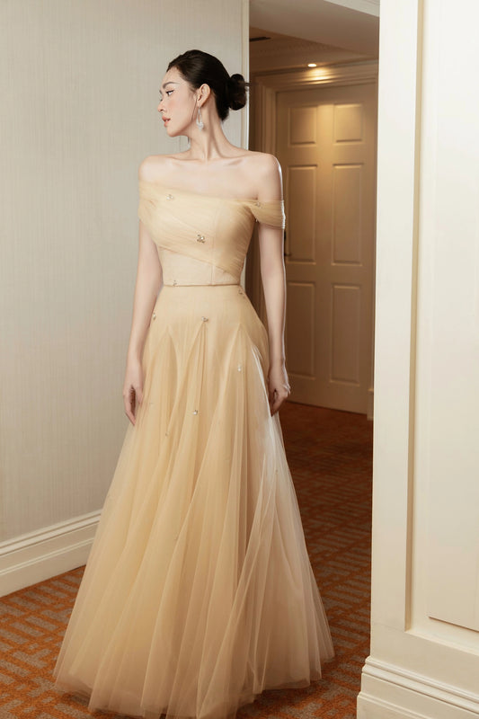 Woman wearing a beige off-shoulder gown standing in a room with neutral walls.