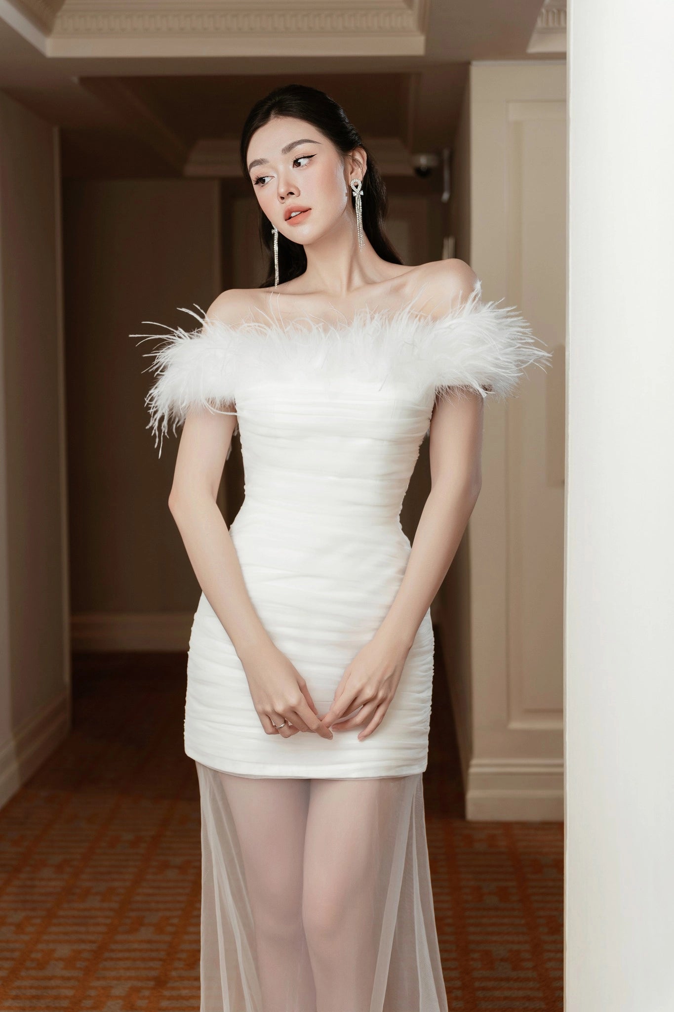 Woman in a white off-shoulder dress with feather details standing indoors.