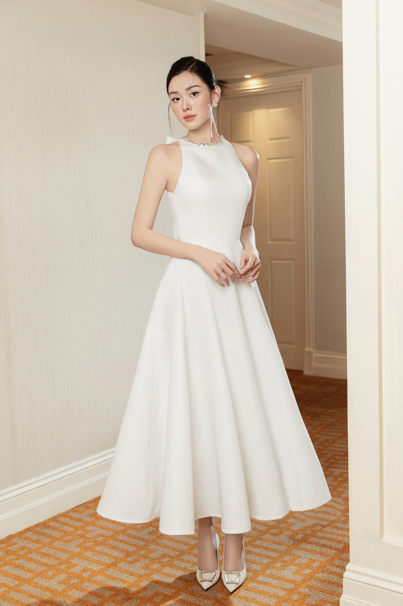 Woman in a white dress standing in an indoor setting with neutral walls.