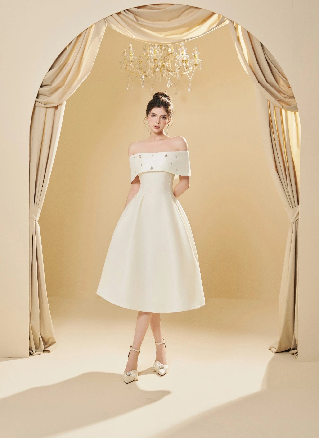 Woman in a white off-shoulder dress standing in front of a decorative archway with curtains.