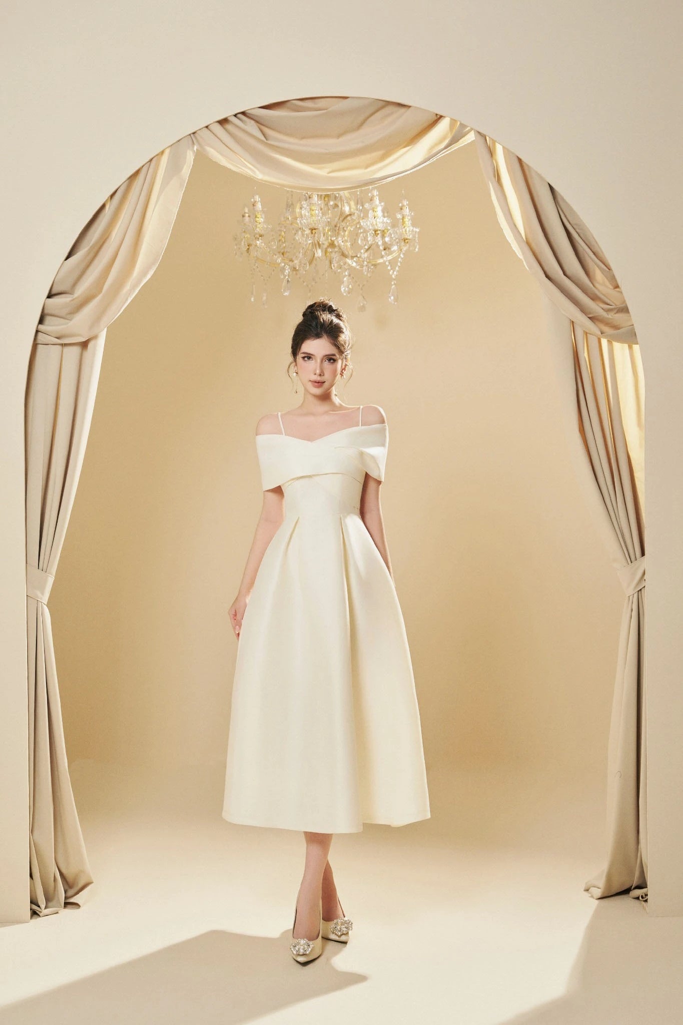 Woman in a white off-shoulder dress standing in front of draped curtains with a chandelier.
