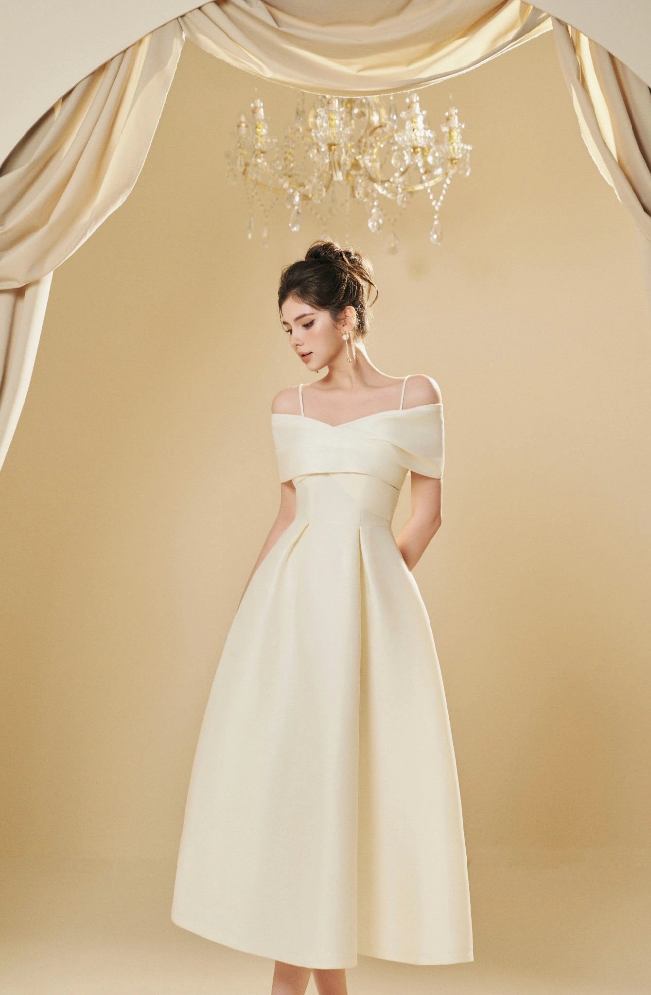 Woman in a white off-shoulder dress standing in front of a chandelier and draped fabric.