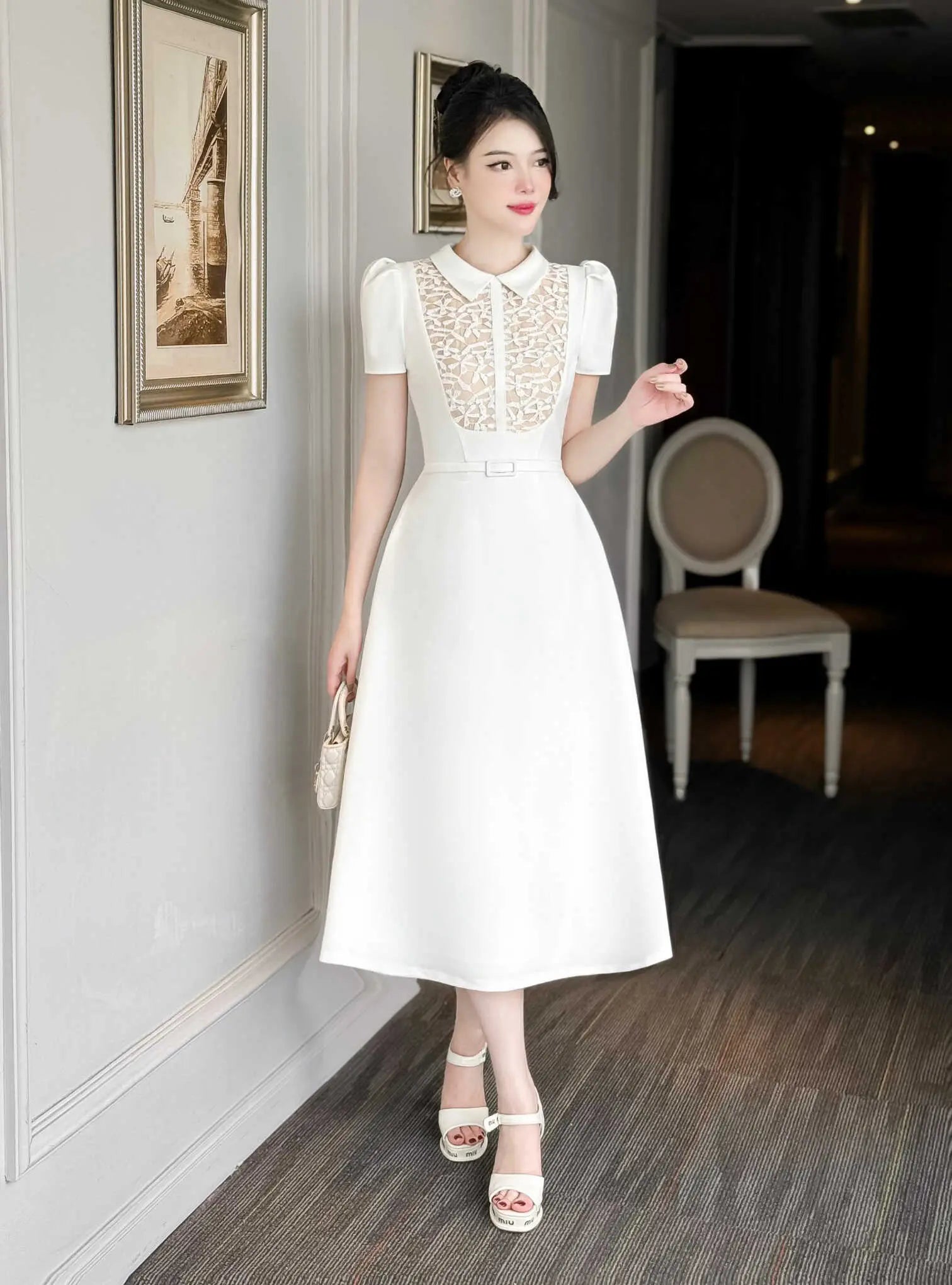 Woman in a white dress standing in an indoor setting with framed pictures on the wall.