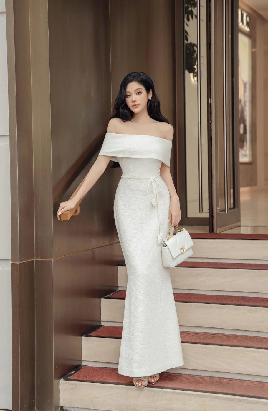 Woman in a white off-shoulder dress standing on steps outside a building.