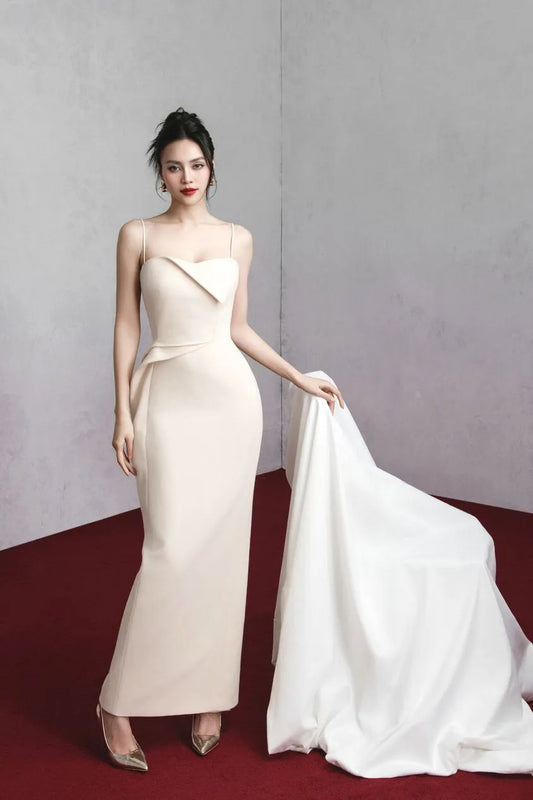 A woman in a cream-colored, sleeveless party dress with a V-neck neckline and spaghetti straps, standing in a graceful pose with one hand behind her head.