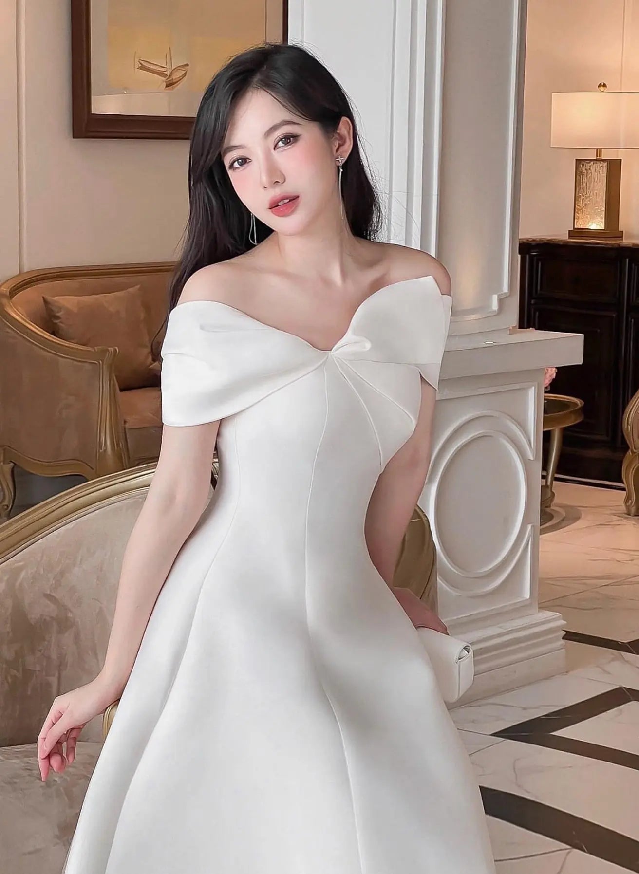 Woman in a white off-shoulder dress standing in an elegant interior setting.