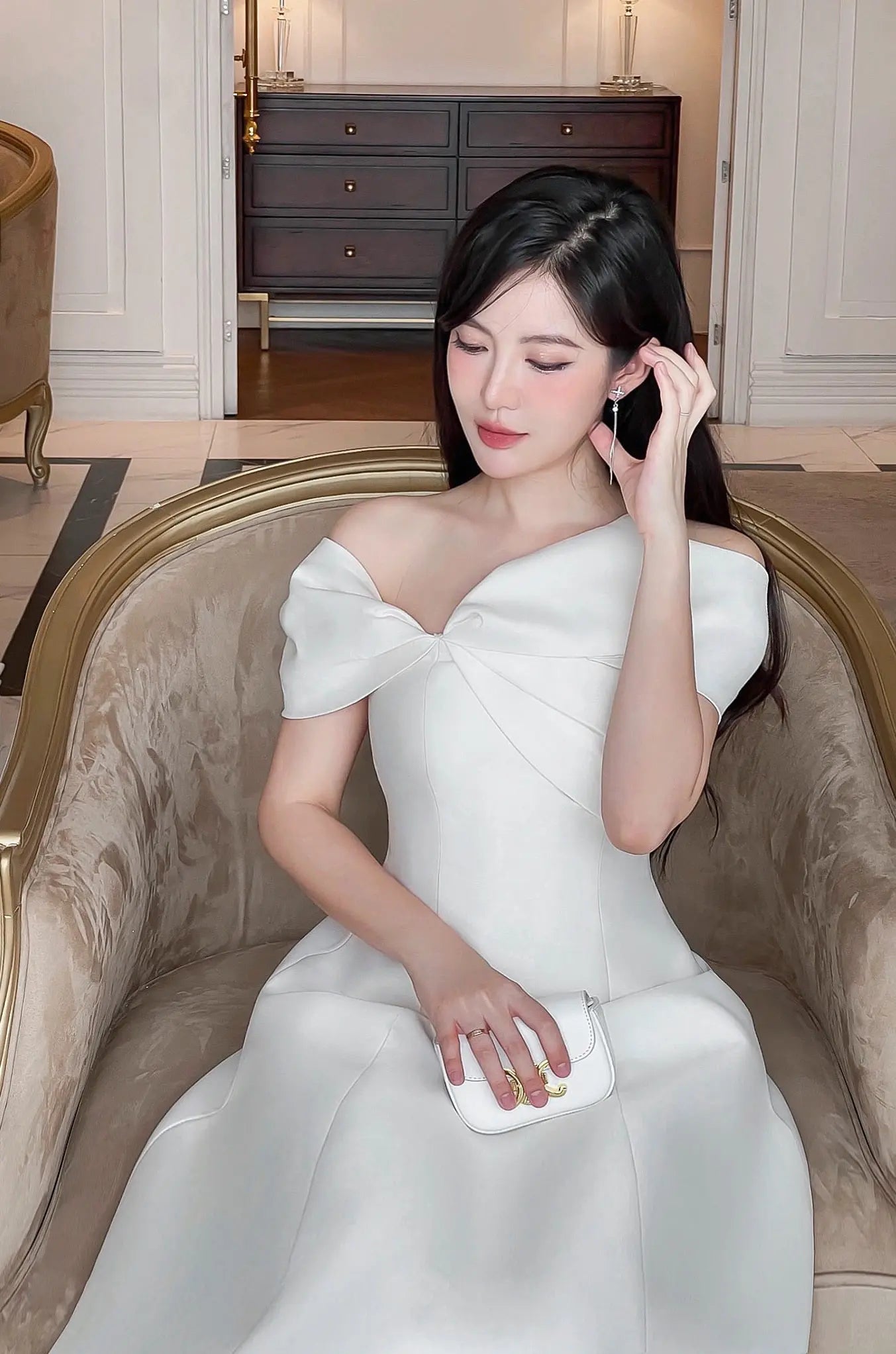 Woman in a white dress sitting on an ornate chair in a luxurious room.