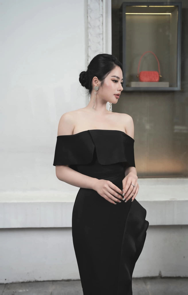 Woman in a black off-shoulder dress standing against a light-colored wall.