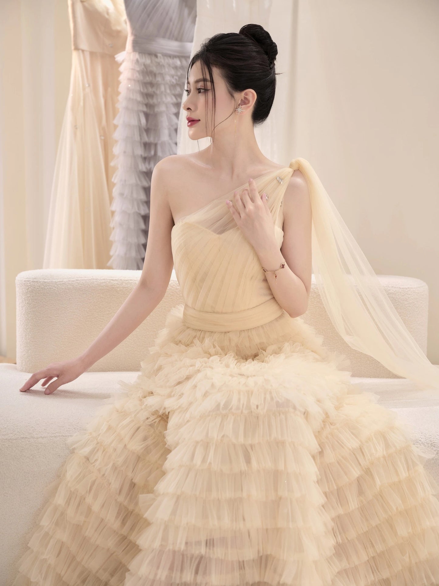 Woman in a light yellow gown sitting on a couch in a room with white walls and a white chair.