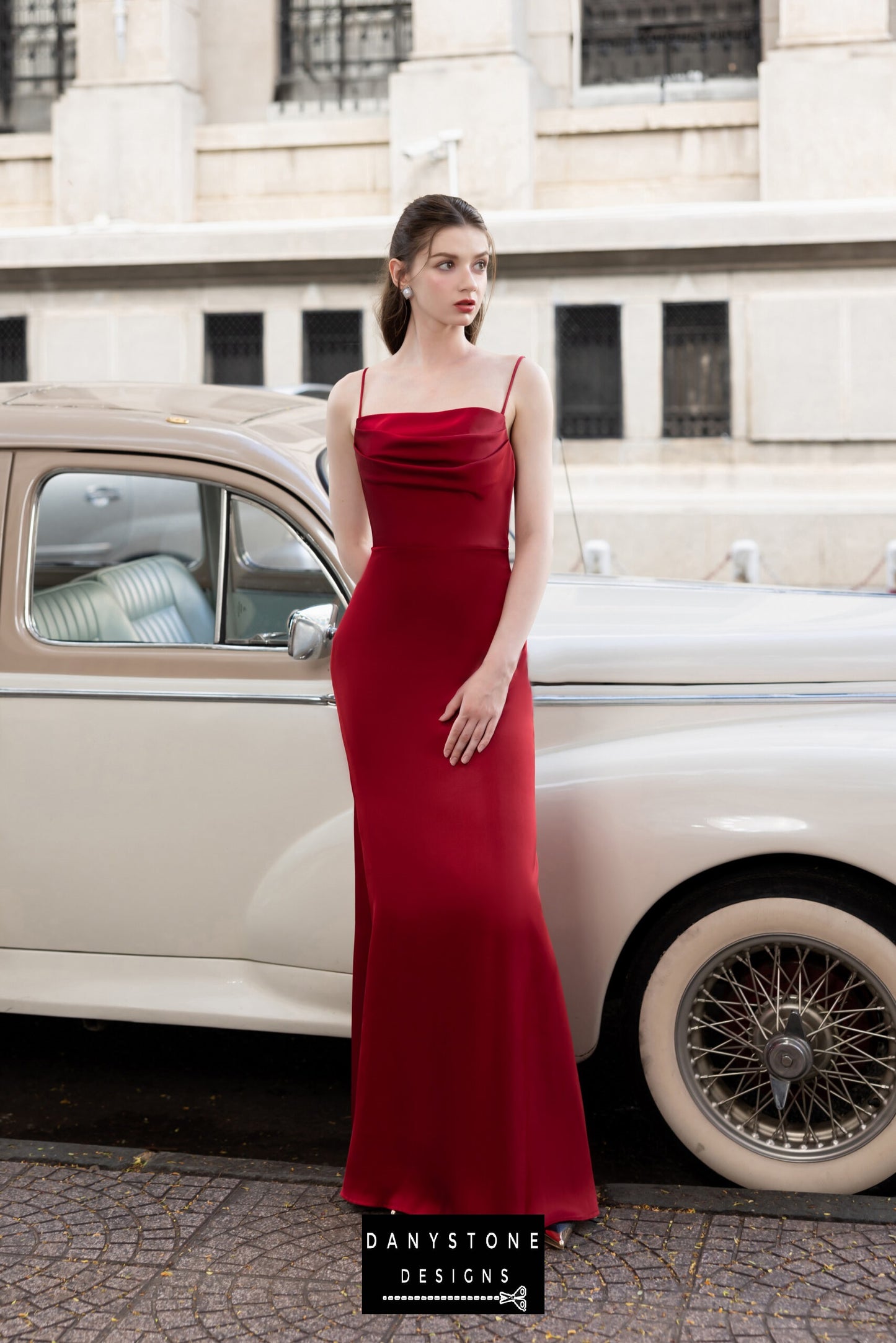 Woman in a luxurious red silk dress with open back and tie detail, standing beside a classic car.