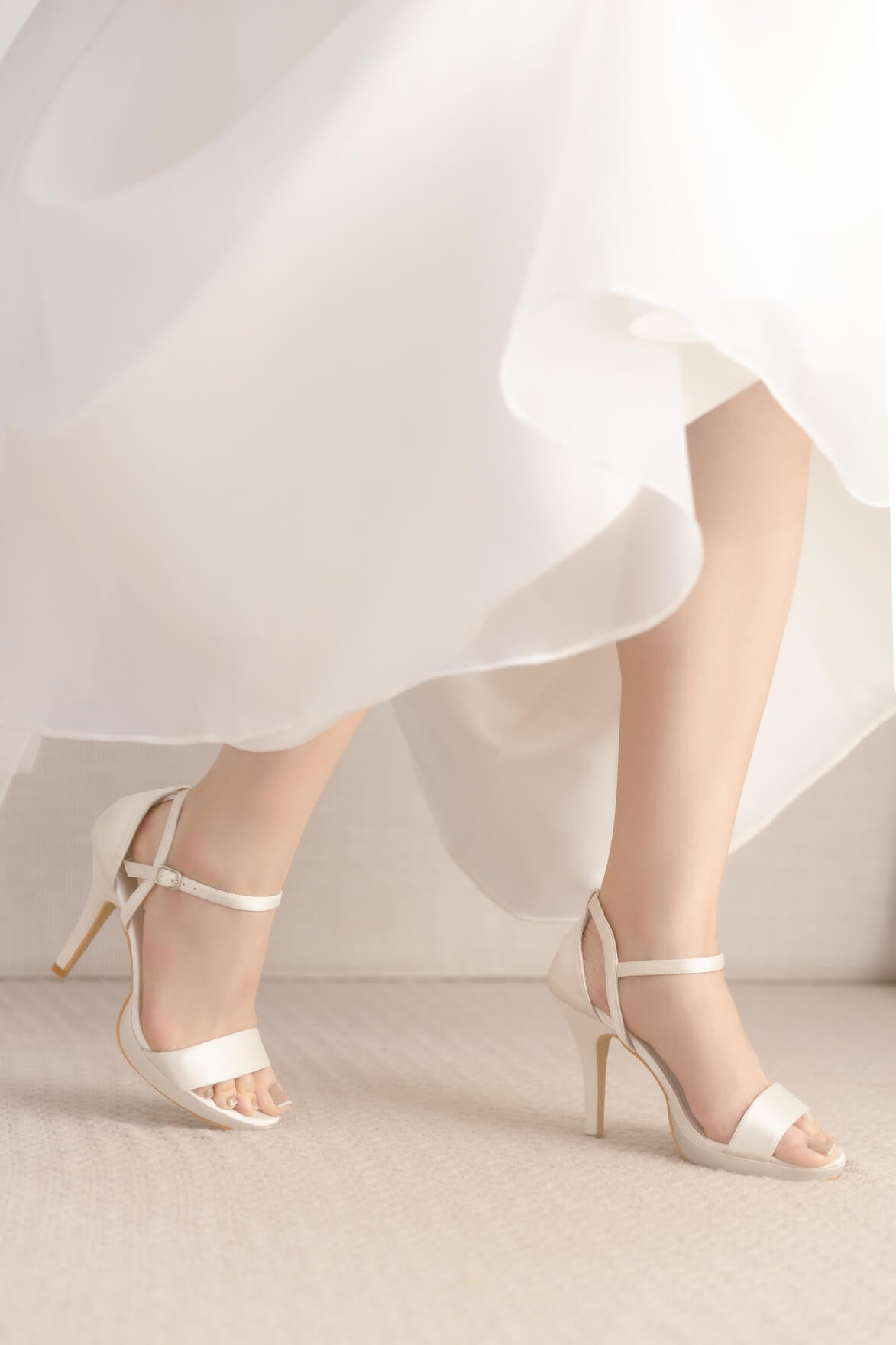 White high-heeled sandals worn with a flowing white dress on a neutral background