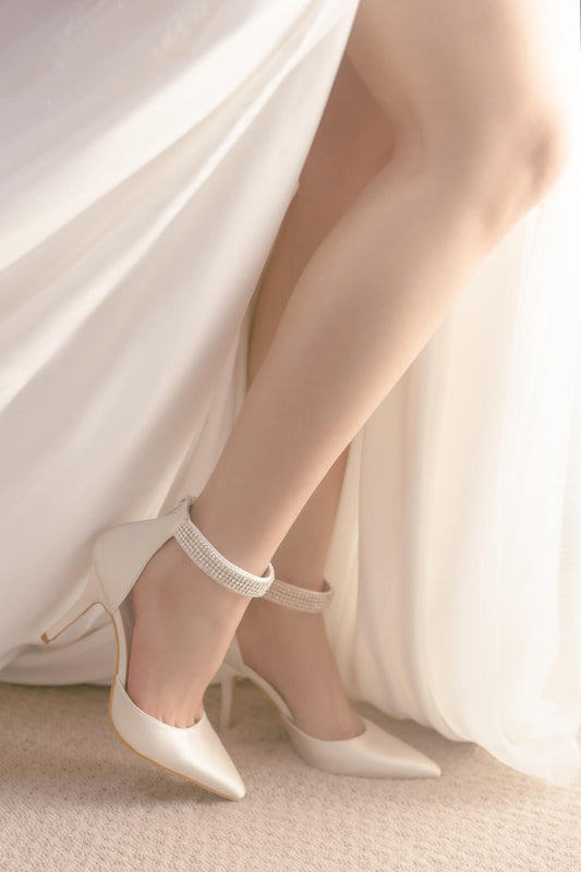 White high-heeled shoes with ankle straps worn by a person in a white dress.