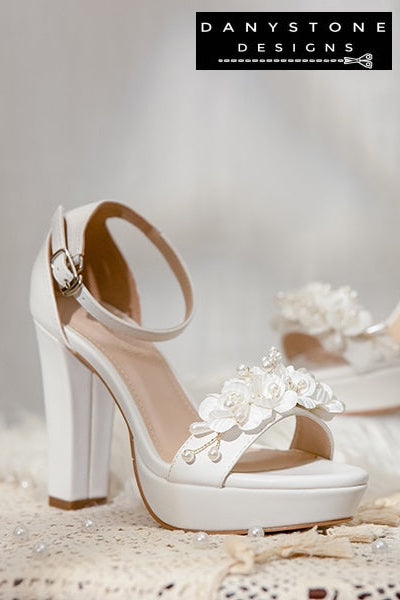 Pair of Elegant White Matte Leather Wedding Sandals with floral embellishments on a lace tablecloth.